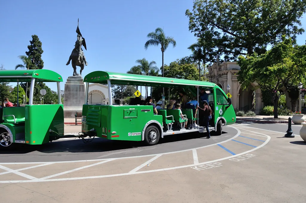 A green open-air Balboa Park tram picking up riders near a statue and palm trees