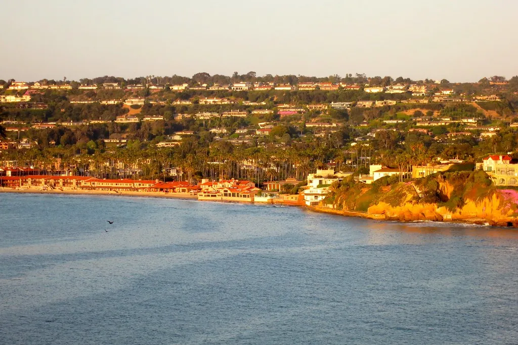 Calm water off La Jolla Shores in warm golden light, with beachfront buildings, palm trees and hillside homes behind the sand