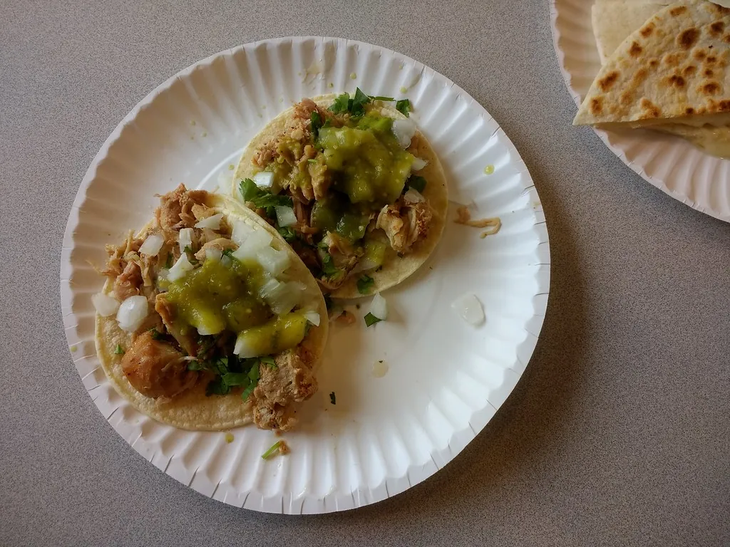 Two shredded meat tacos with green salsa, onion and cilantro on a paper plate