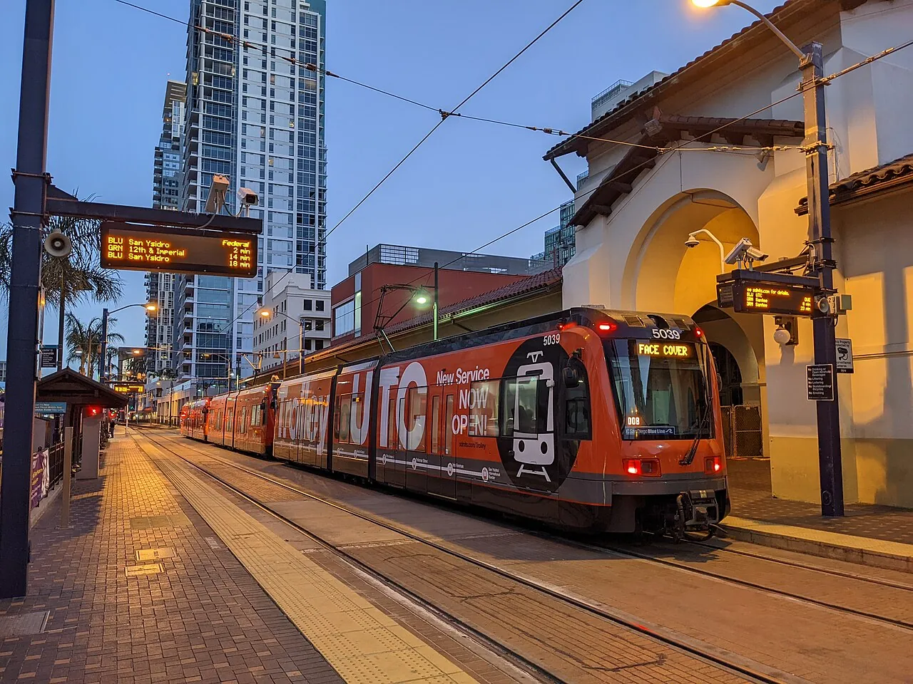 A UC San Diego Blue Line trolley waiting at the Santa Fe Depot station at dusk, with downtown towers behind